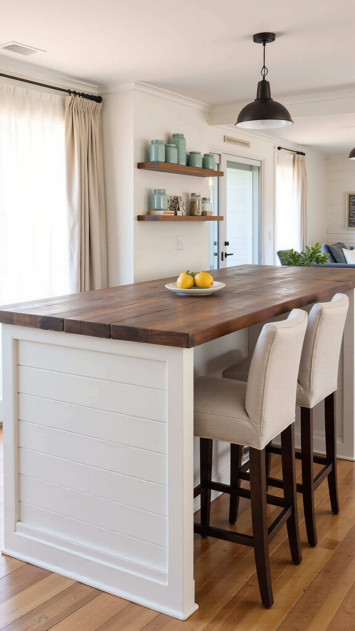 Open-concept kitchen at golden hour with L-shaped white shiplap island, dark walnut countertop, oatmeal linen barstools, floating shelves with vintage decor, and ambient black sconces.