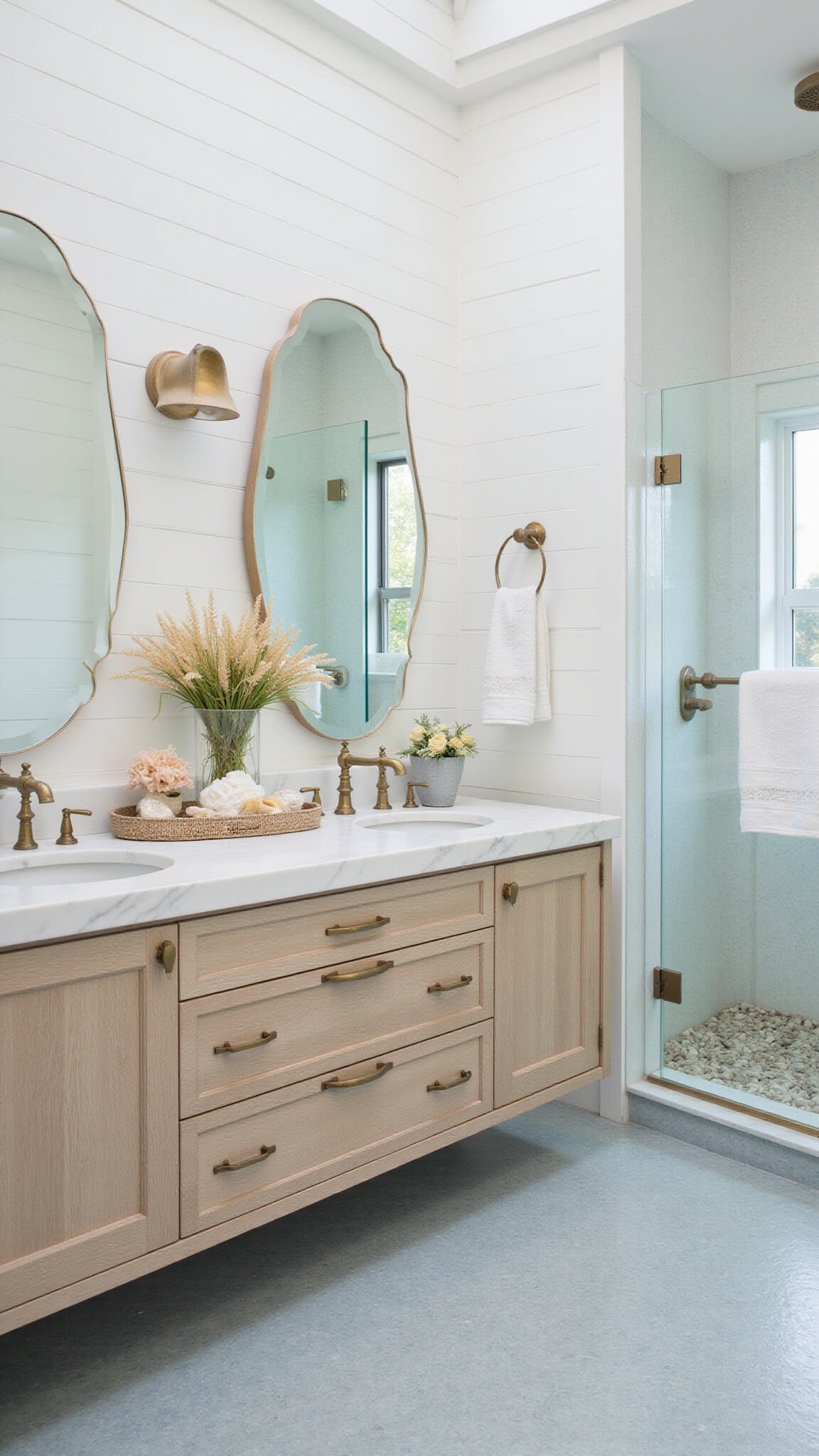 Modern coastal bathroom with white shiplap walls, pale blue penny tile floor, floating bleached oak double vanity, brass hardware, round mirrors, glass shower, and coastal decor in bright morning light.