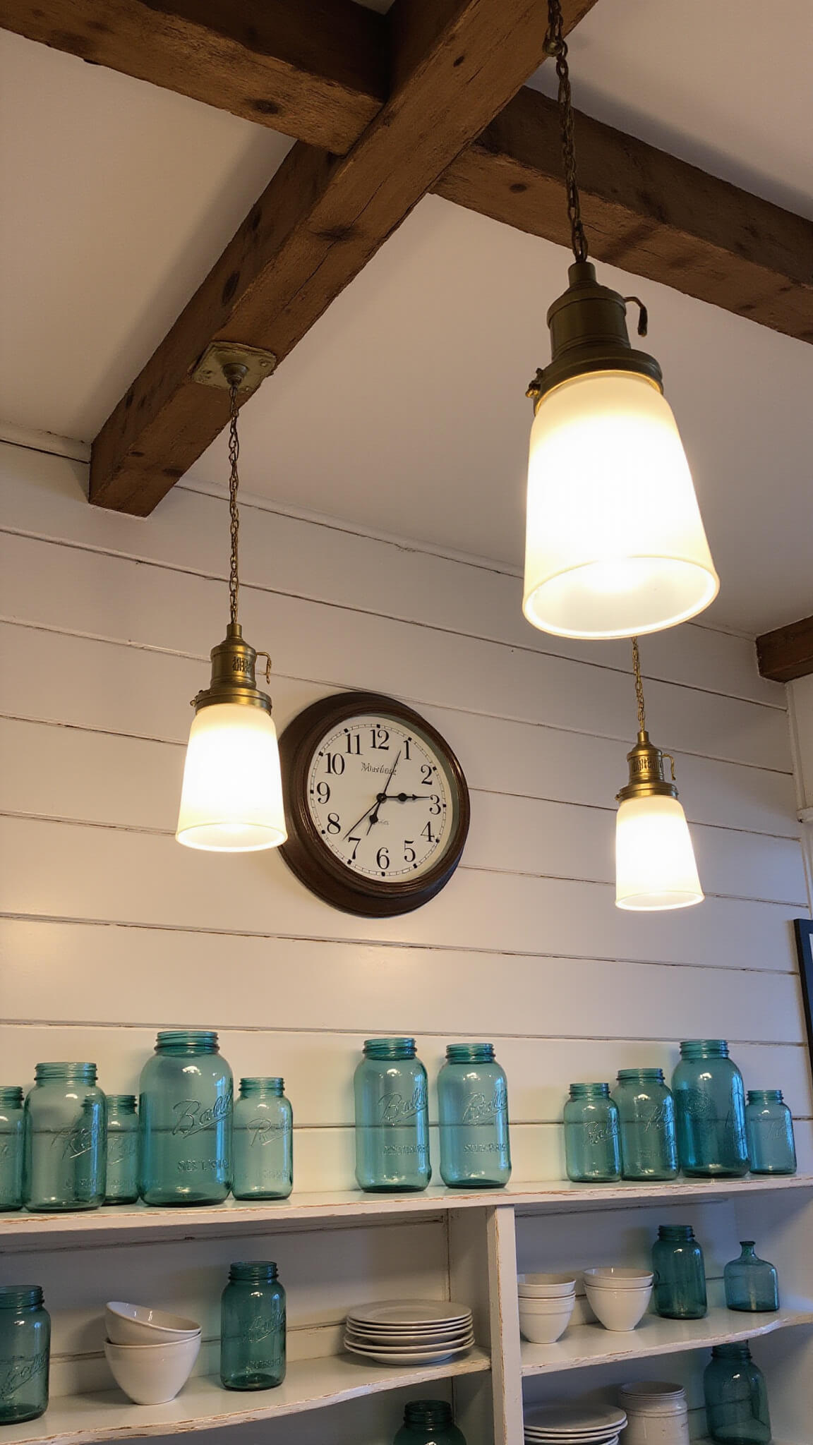 Cozy evening kitchen with brass pendant lights, open shelves displaying blue mason jars and white ceramics, distressed wood ceiling beams, and an antique clock reading 6:45 on a shiplap wall.