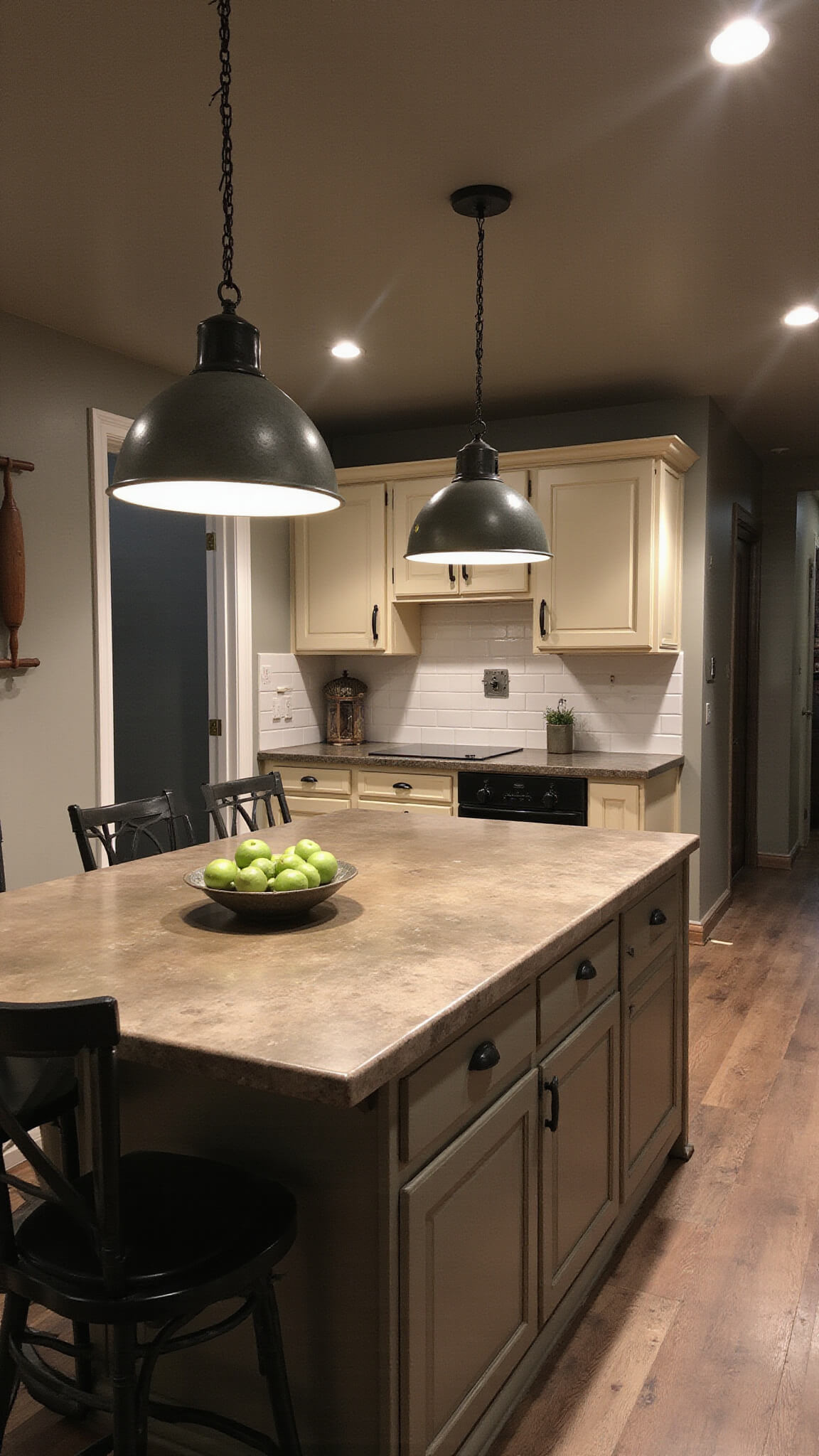 Dramatic, rustic open-concept kitchen at dusk with warm zinc pendant lights over soapstone island, antique rolling pins on wall, and cream cabinets with black iron hardware.