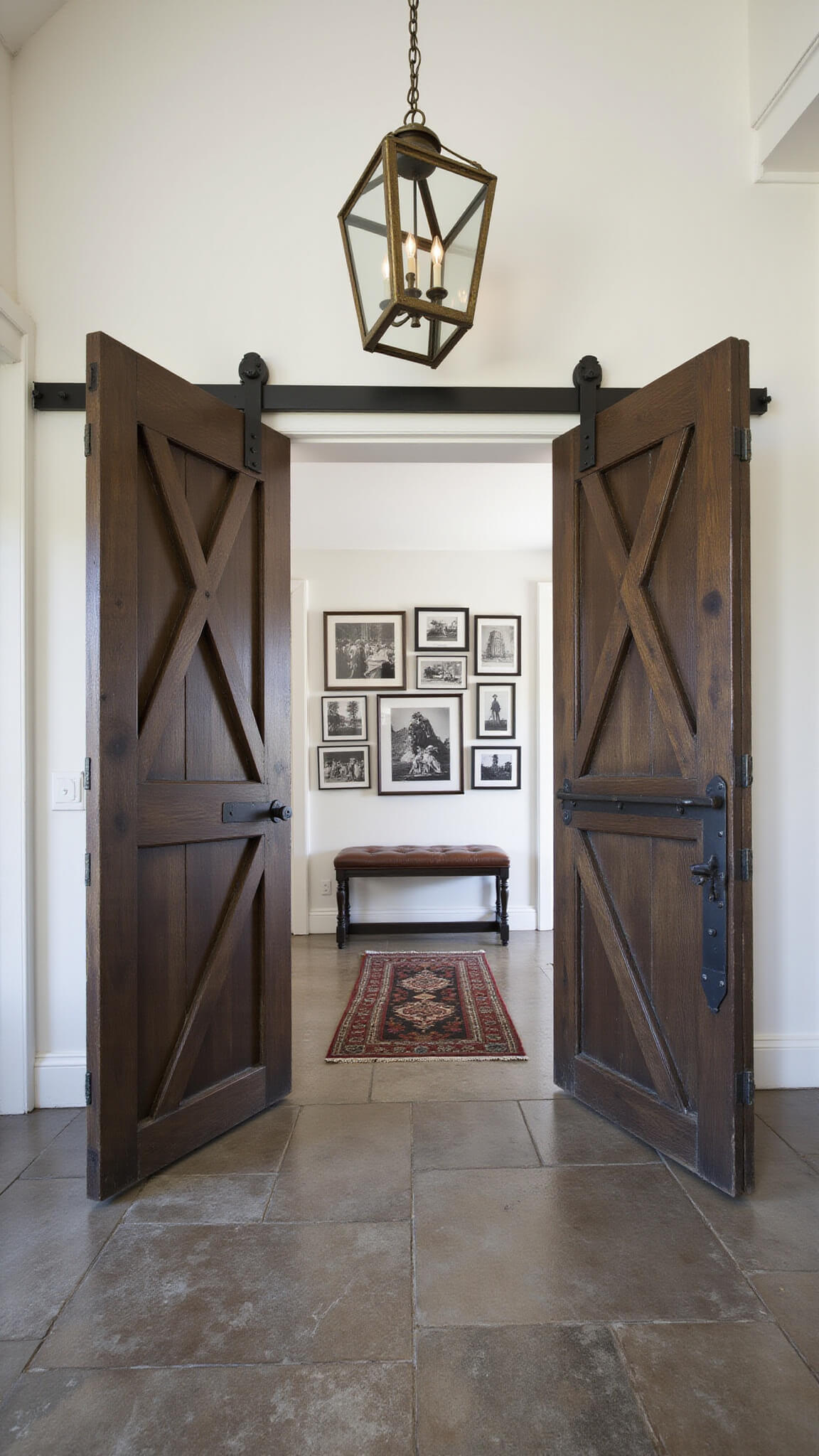 Partially open restored barn doors reveal a dramatic double-height foyer with slate flooring, antique bench, brass chandelier, and black-and-white photo gallery.