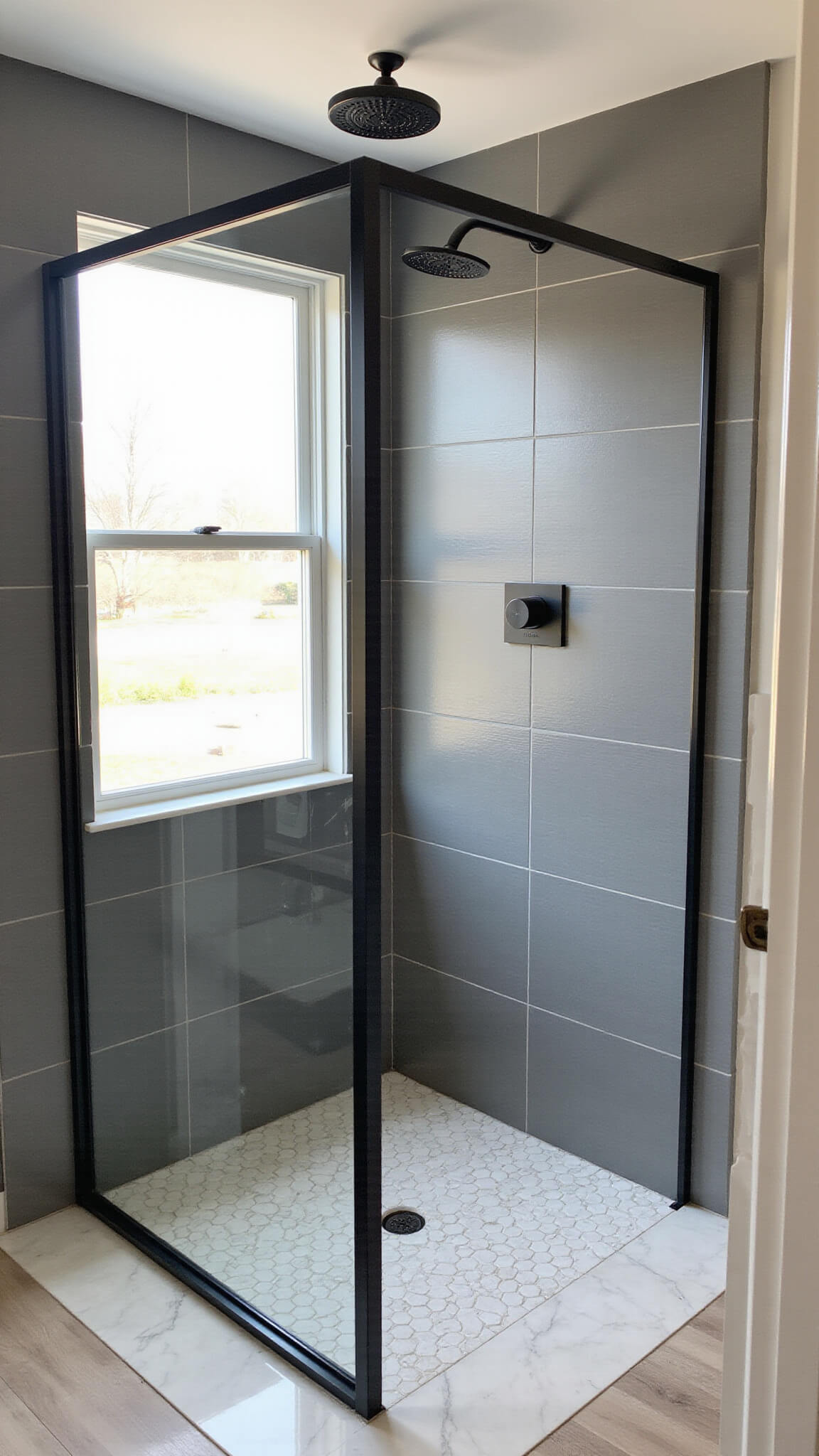 Modern minimalist bathroom with corner doorless shower, charcoal porcelain tiles, white marble hexagon floor, black fixtures, and morning light streaming through a frosted window.