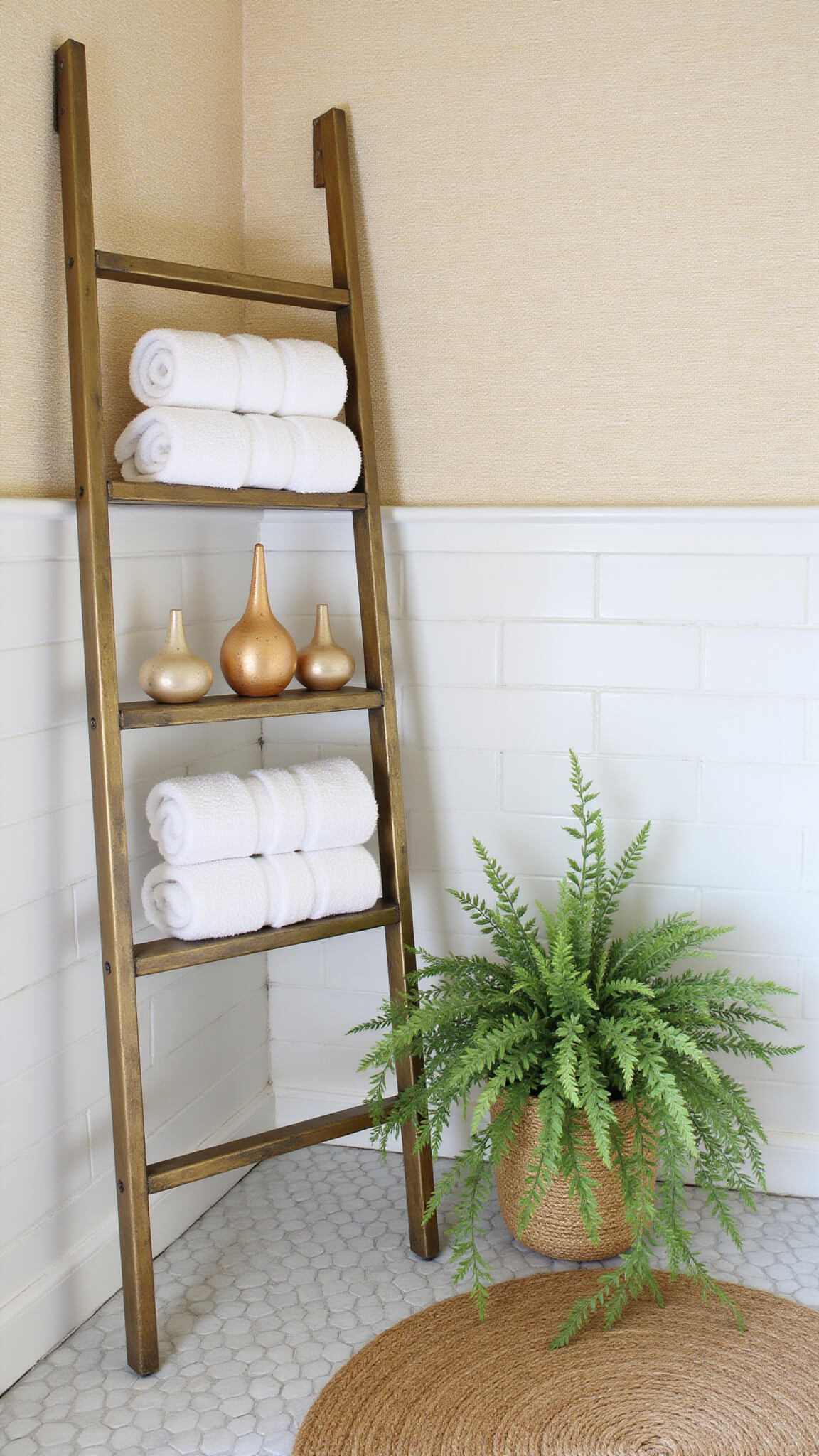 Compact bathroom corner with antique brass ladder shelf holding towels, ceramics, and trailing plant; white subway tiles contrast with cream grasscloth wallpaper and jute rug on hex tile floor.