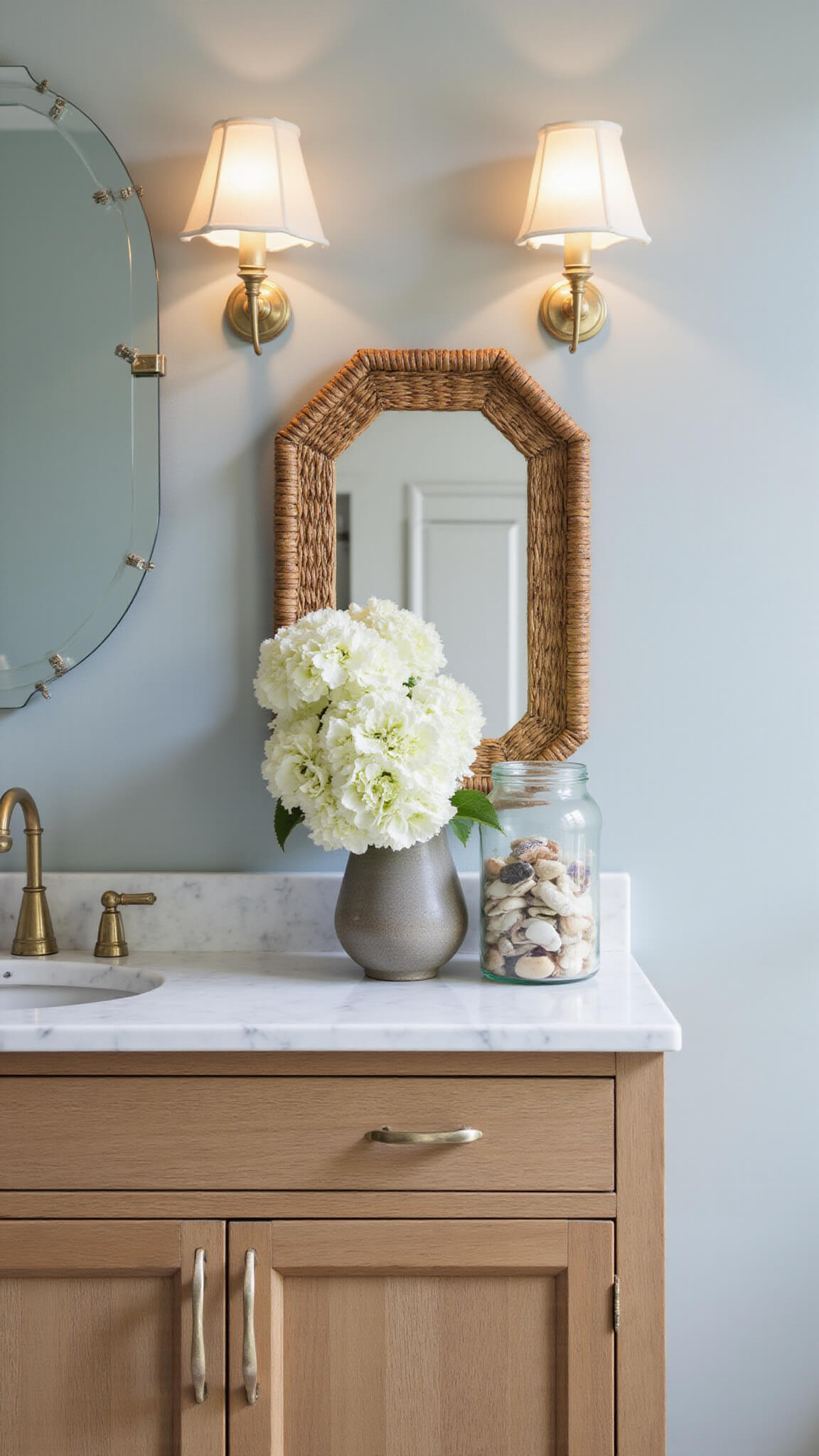 Closeup of white oak floating vanity with Carrara marble top in soft morning light, glass sconces glowing against pale blue-gray walls, vintage apothecary jars, rattan mirror, and white hydrangeas in ceramic vase.