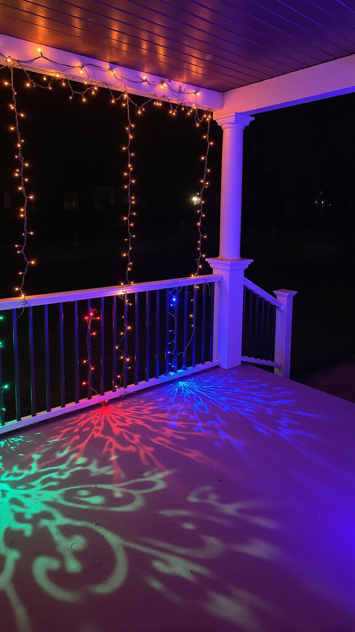 Nighttime view of 15'x15' wraparound porch corner with orange and purple string lights forming a canopy, LED spotlights casting creature-like shadows through props, and color-changing bulbs highlighting architectural details.