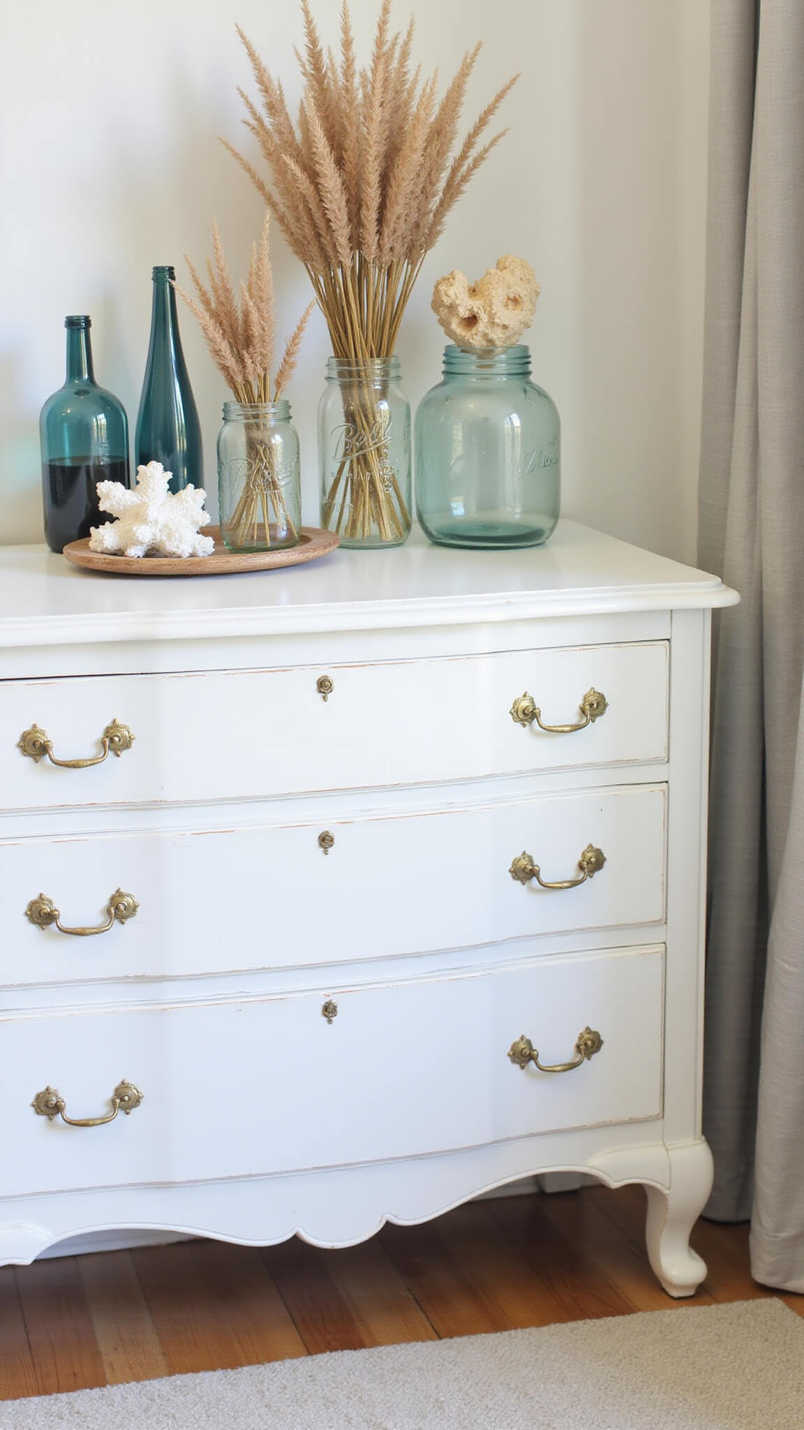 Vintage coastal farmhouse dresser with brass pulls, styled with mason jars of dried beach grass, coral pieces, and weathered blue glass bottles, softly lit by morning side-light with a blurred background.