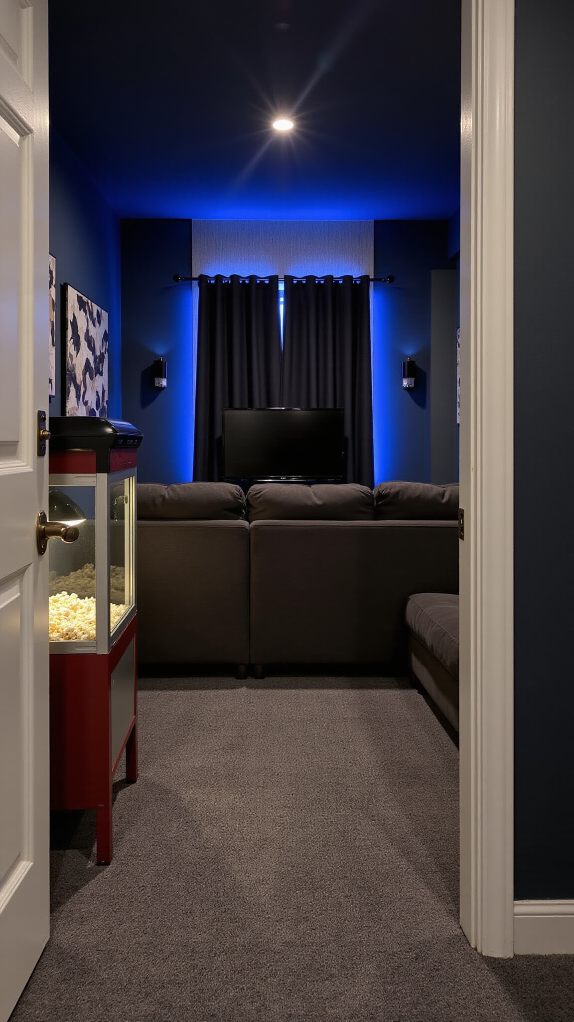 Cozy 10'x12' home theater room with dark navy walls, plush charcoal sectional, 65" OLED TV, ambient blue LED lighting, theater sconces, acoustic panels, and popcorn machine.