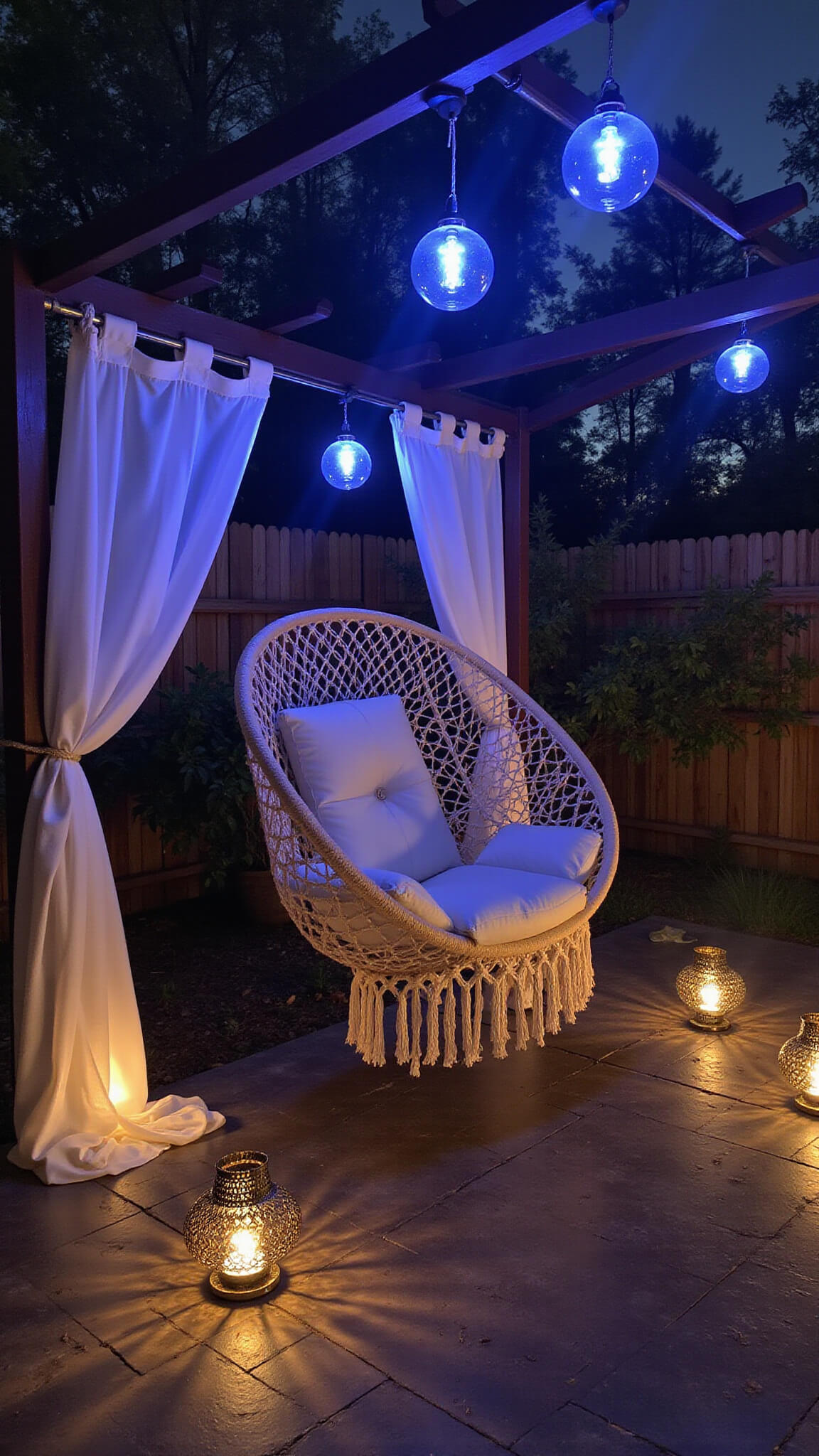 Mystical dusk lounge with macramé hanging chair under pergola, gauzy curtains, blue-lit glass globes, and Moroccan lanterns casting patterns on stone floor.
