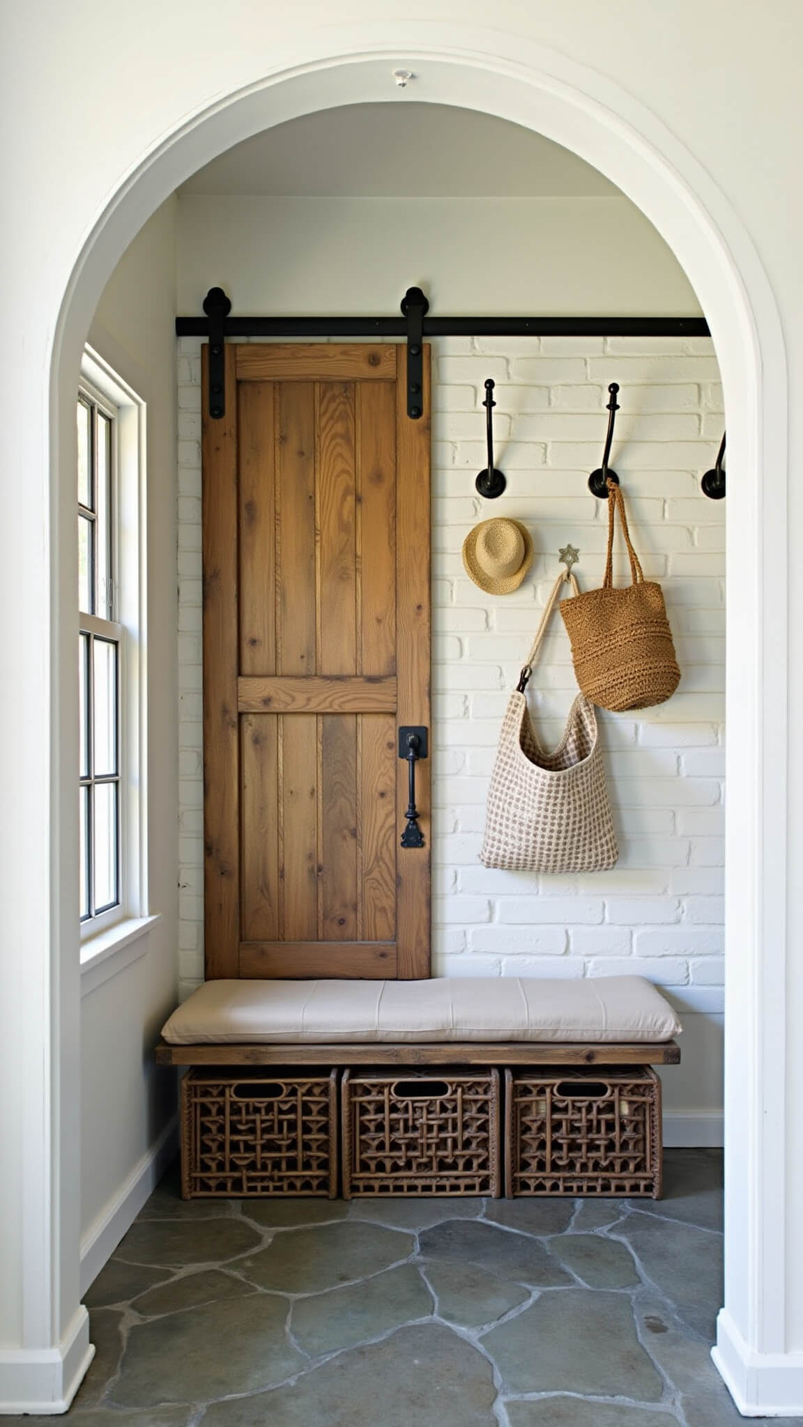 Cozy entryway nook with arched barn door, reclaimed wood bench over vintage crates, whitewashed brick wall, straw hats, and market bags, lit by soft morning light.