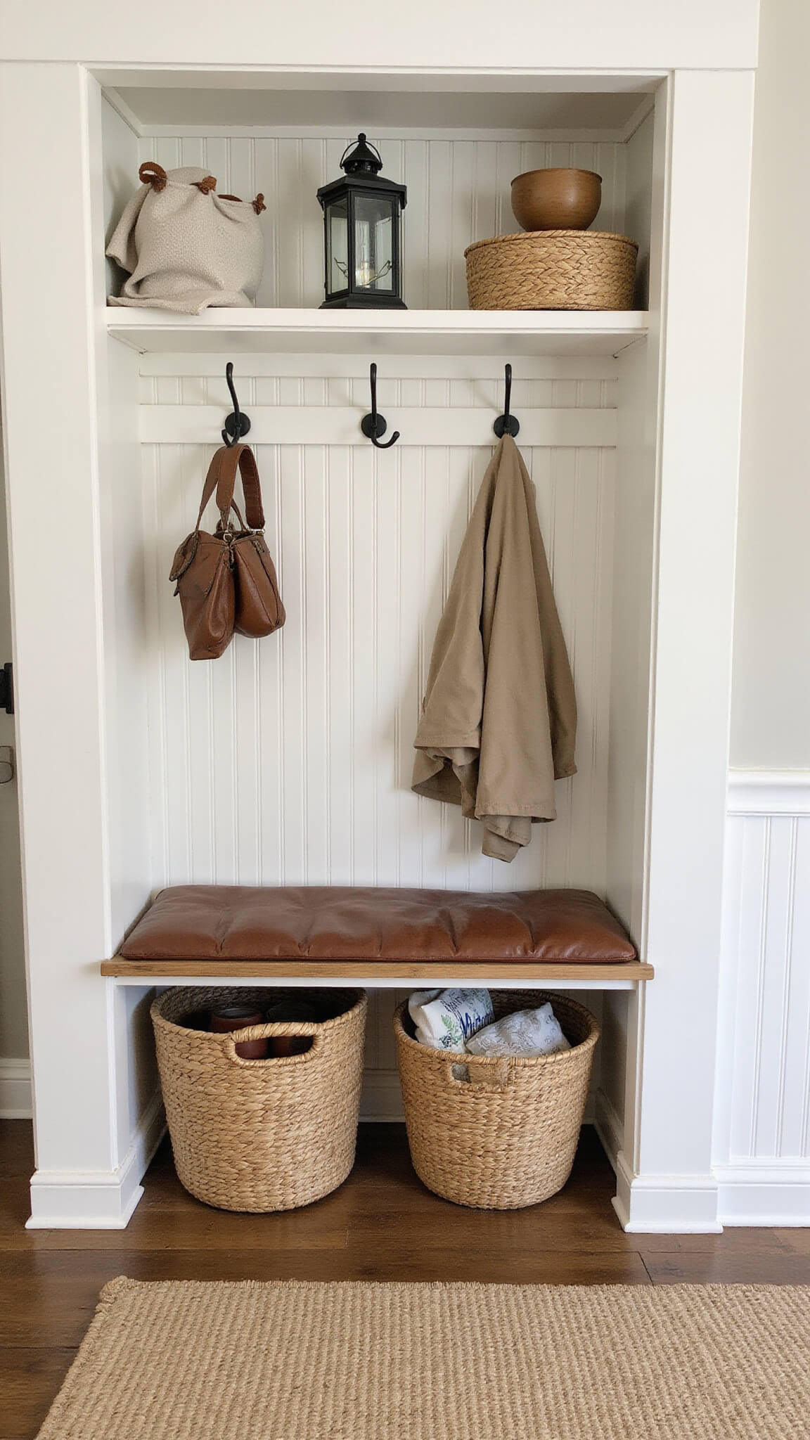 Mudroom with built-in bench, worn leather cushion, vintage hooks holding outerwear, reclaimed timber floors, white beadboard walls, woven baskets, and vintage lantern for lighting.