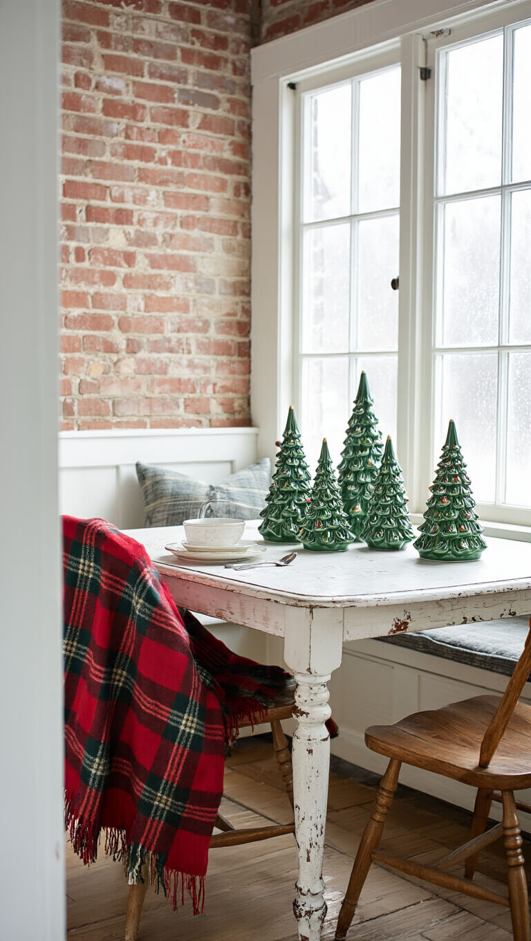 Cozy breakfast nook with vintage 1960s ceramic Christmas trees on distressed farmhouse table, plaid blanket on Windsor chair, exposed brick wall, and frosty window light.