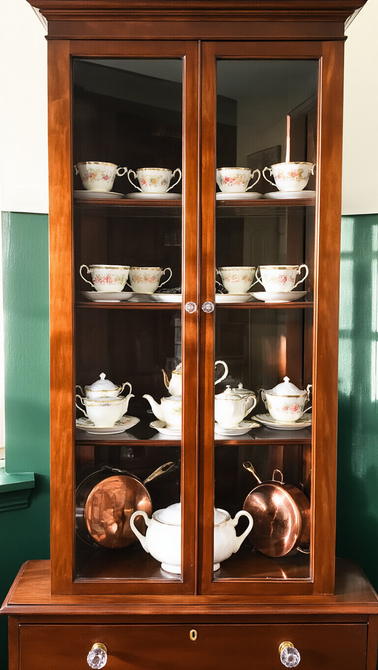 Victorian cherry wood display cabinet with bone china teacups on glass shelves, softly lit against emerald walls with sparkling crystal knobs and warm copper reflections.