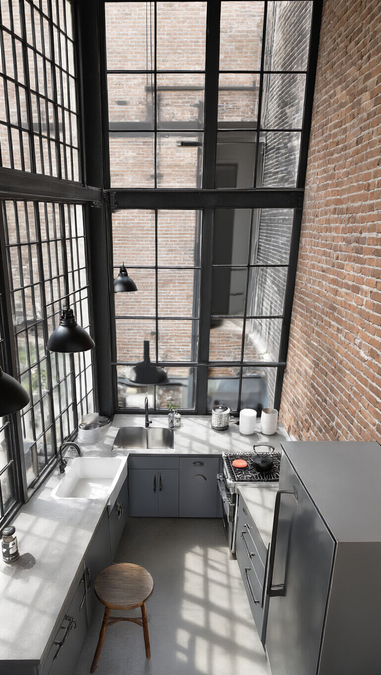 Overhead view of industrial chic gray kitchen with steel cabinets, concrete countertops, black-framed windows, exposed brick wall, and industrial lighting.