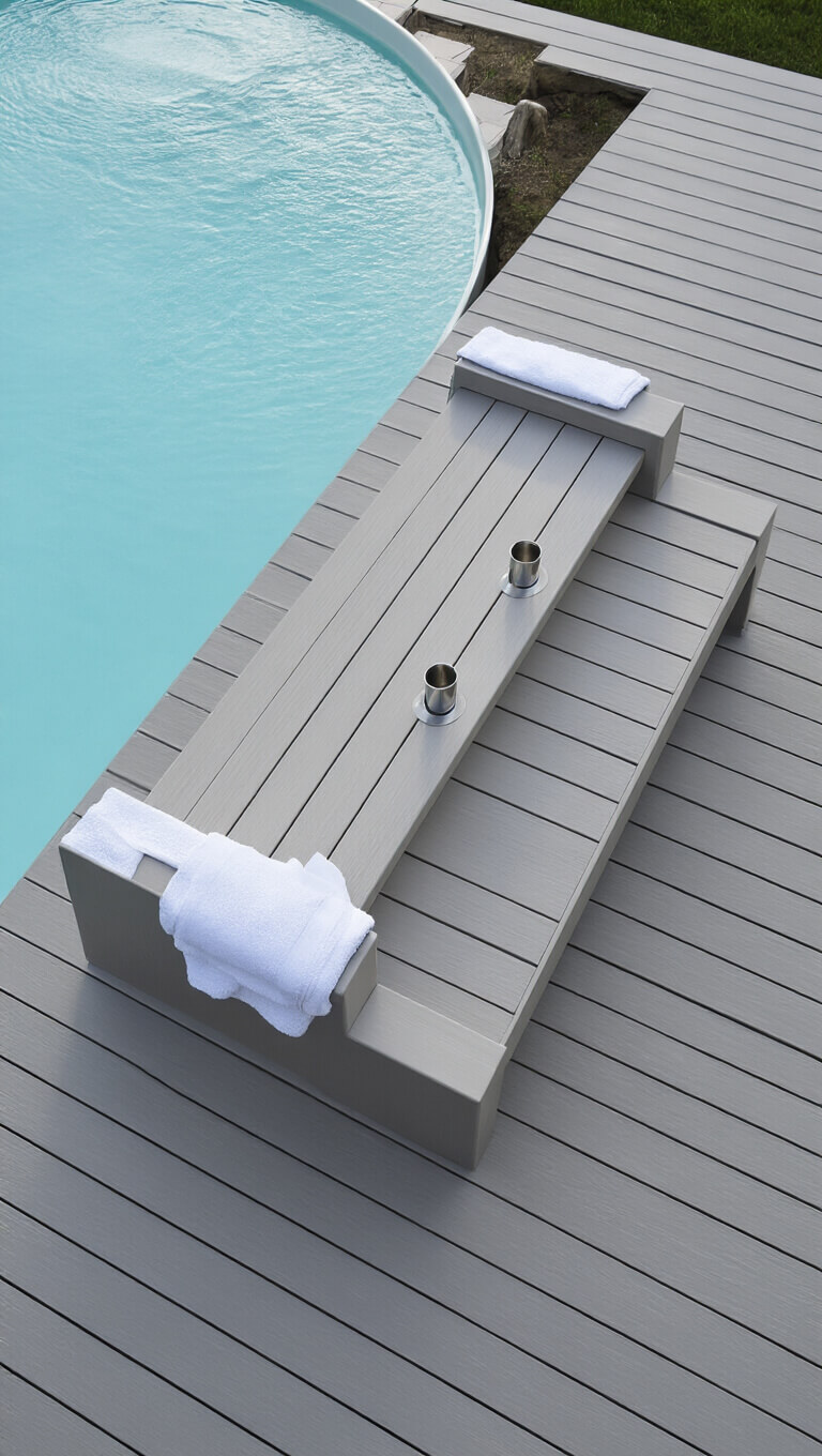 Minimalist backyard with stock tank pool centered on light grey composite decking bench; early morning light highlights clean lines, chrome drink holders, and draped white towels.
