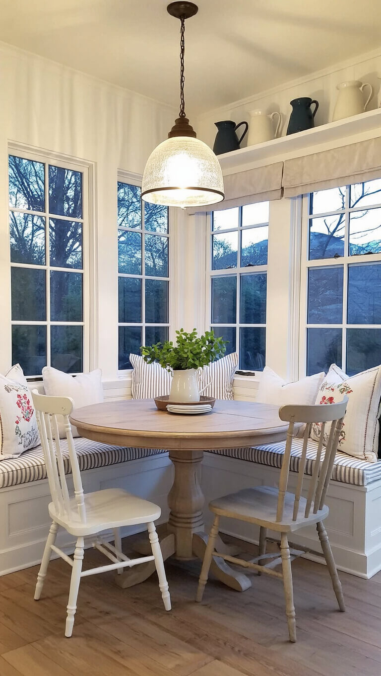 Cozy twilight dining nook with round oak table, mismatched white chairs, striped window seat cushions, ironstone pitchers on shelves, and warm pendant light.